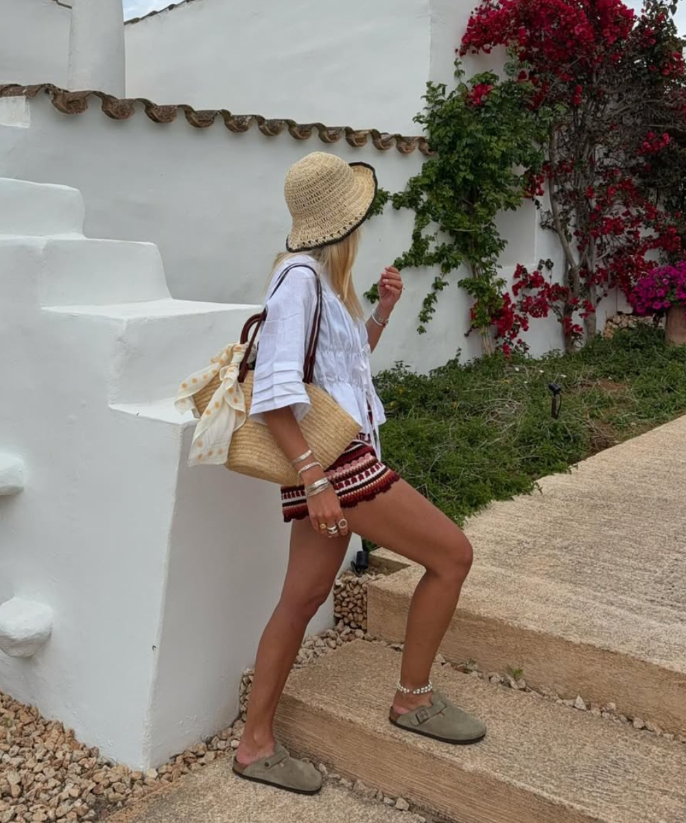 Modelo posa em short de crochê vermelho, branco e marrom com blusa branca ampla, chapéu e bolsa de palha, em clima leve e