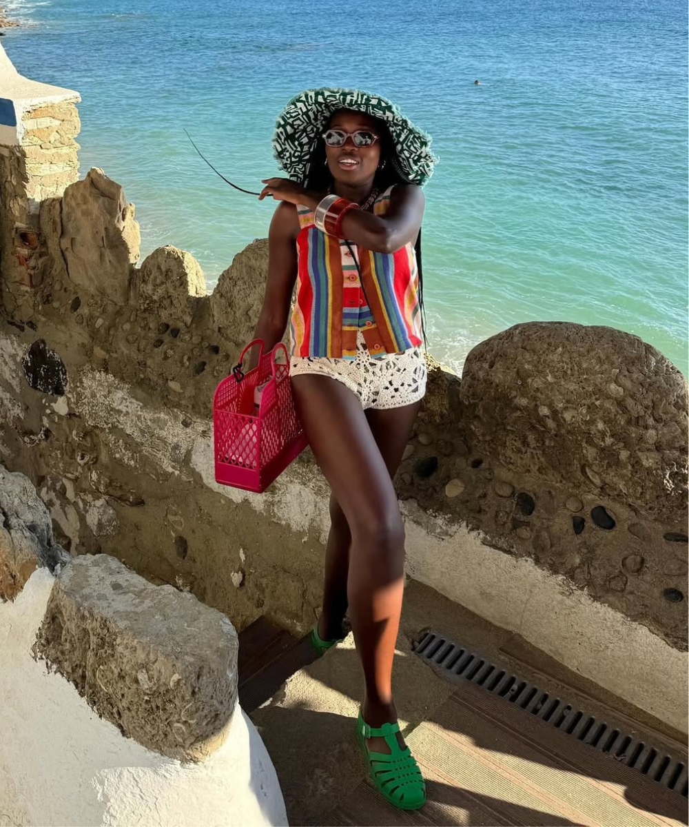 Modelo posa à beira-mar com short de crochê branco, blusa listrada colorida, chapéu estampado e acessórios vibrantes.