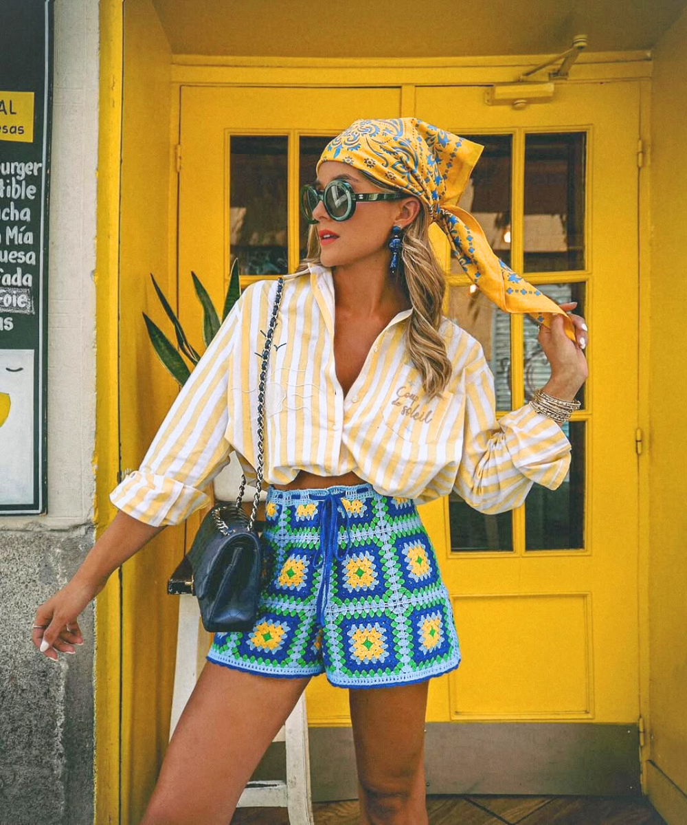 Modelo posa com short de crochê colorido em azul, verde e amarelo, combinando com camisa listrada amarela e lenço vibrante.
