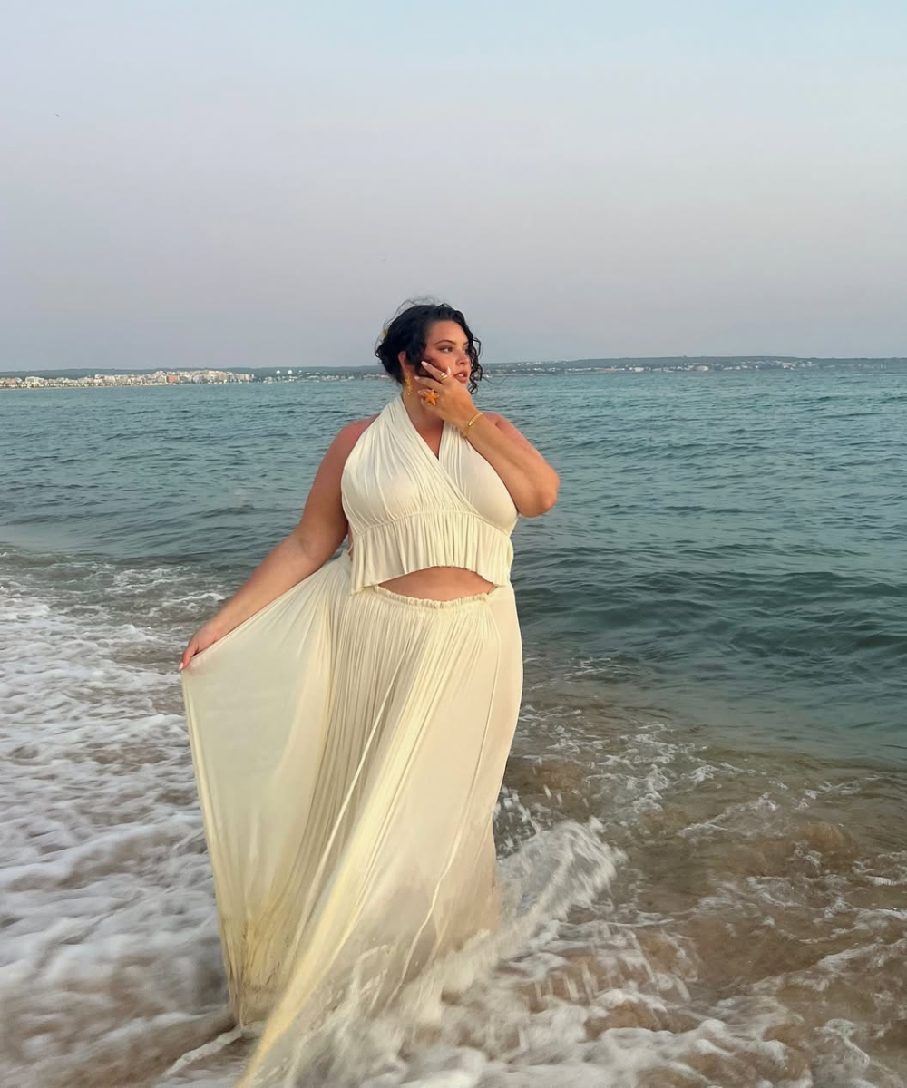 Modelo posa à beira-mar com conjunto estiloso em tecido fluido off-white, top halter e saia longa plissada que realçam