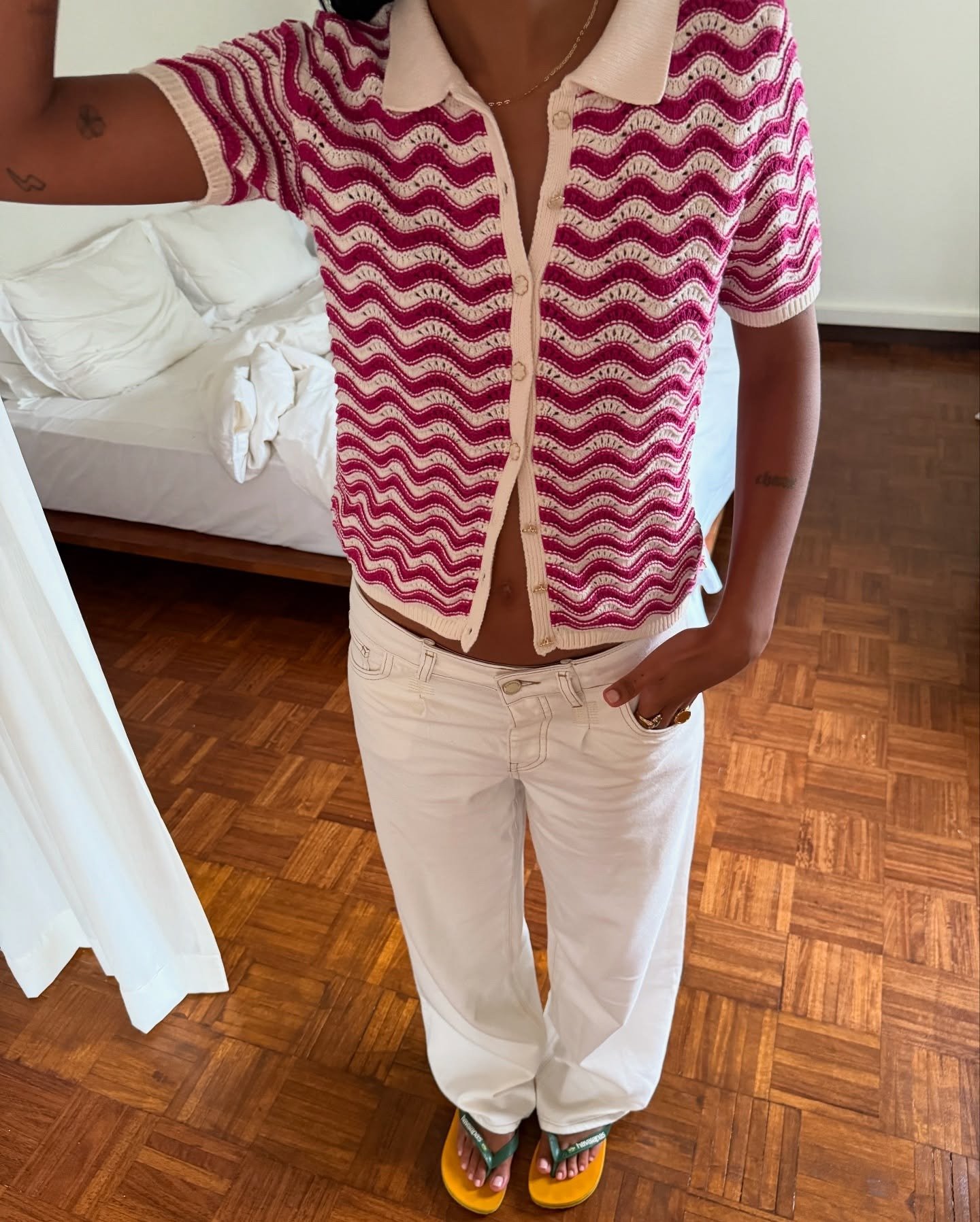 A imagem apresenta uma pessoa usando um cardigã de crochê com padrão ondulado em tons de rosa e um colar delicado. A parte inferior é composta por calças brancas largas. Nos pés, destacam-se chinelos Havaianas em cores vibrantes: verde e amarelo.