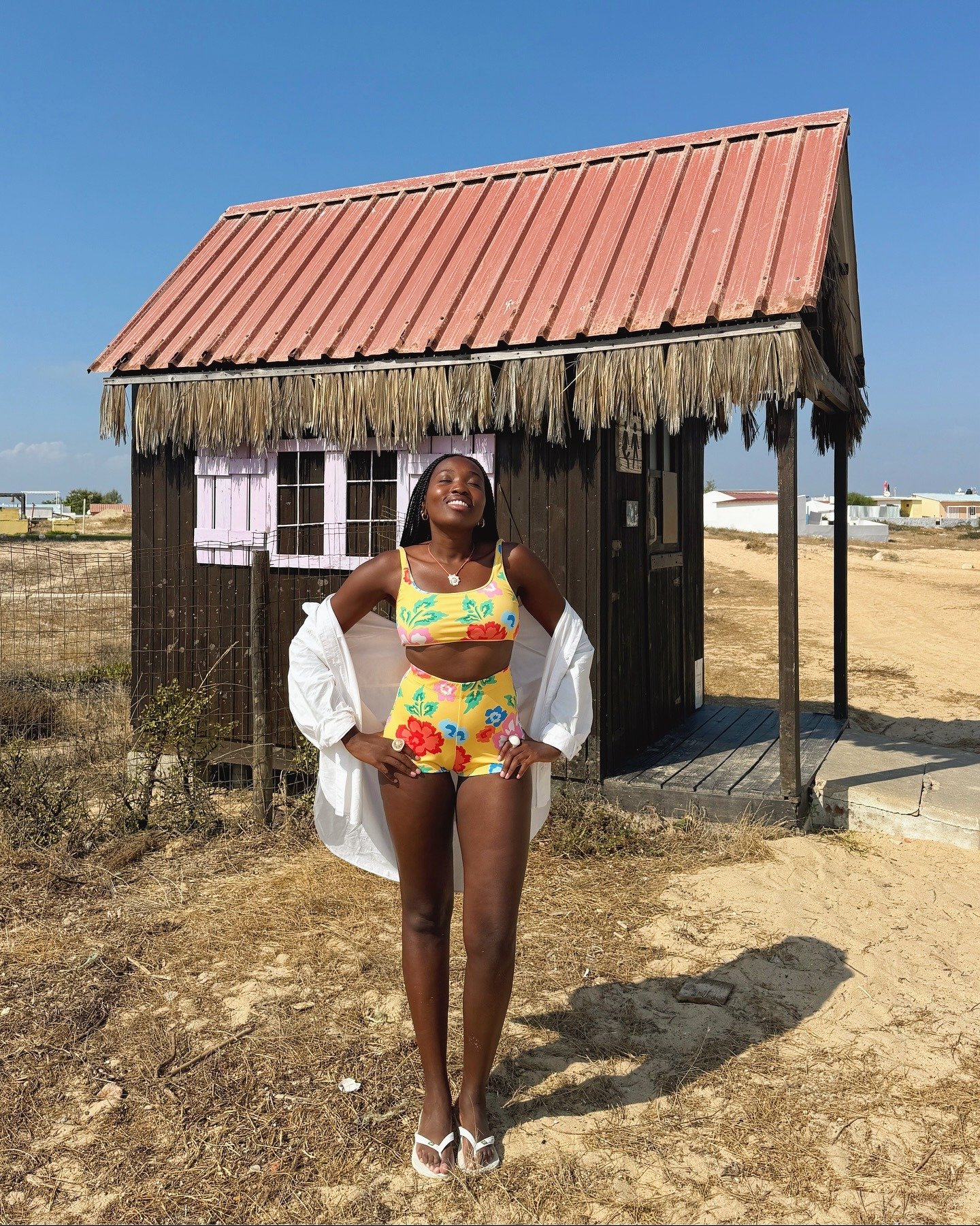 A imagem apresenta uma mulher sorridente, vestindo um biquíni colorido com estampas florais vibrantes em amarelo, azul e rosa. Ela usa uma camisa branca leve, ideal para o calor.