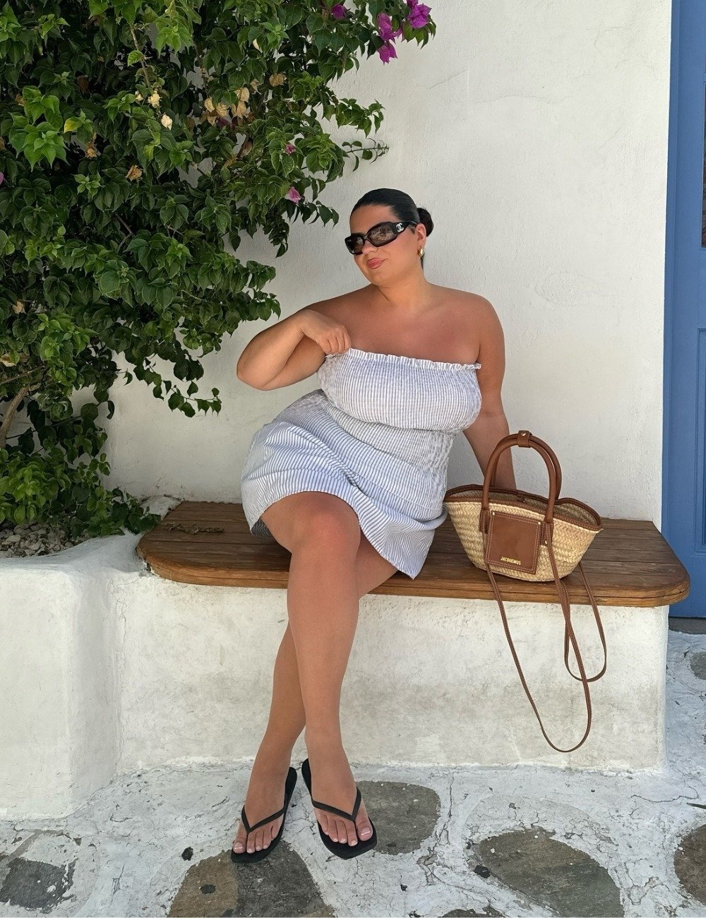 A imagem apresenta uma mulher sentada em um banco de madeira, vestindo um vestido listrado e chinelos Havaianas pretos. Ela combina o look com óculos escuros e uma bolsa de palha, transmitindo uma estética casual e elegante.