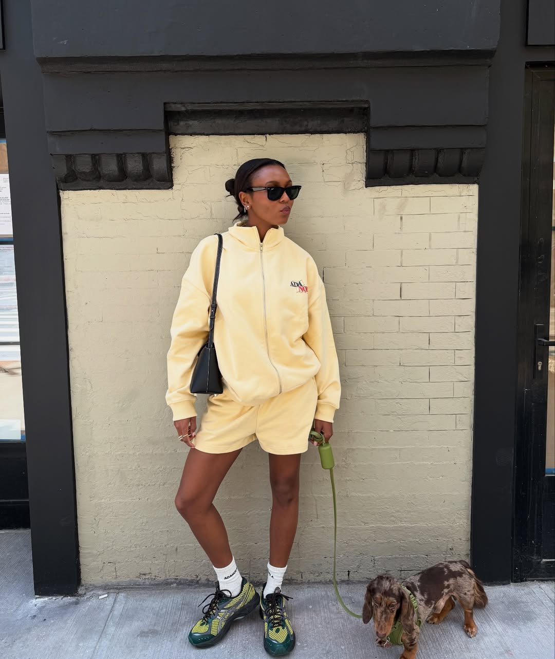 Mulher com conjunto amarelo, cor de Leão para 2026, posa com óculos escuros enquanto passeia com cão salsicha na calçada.