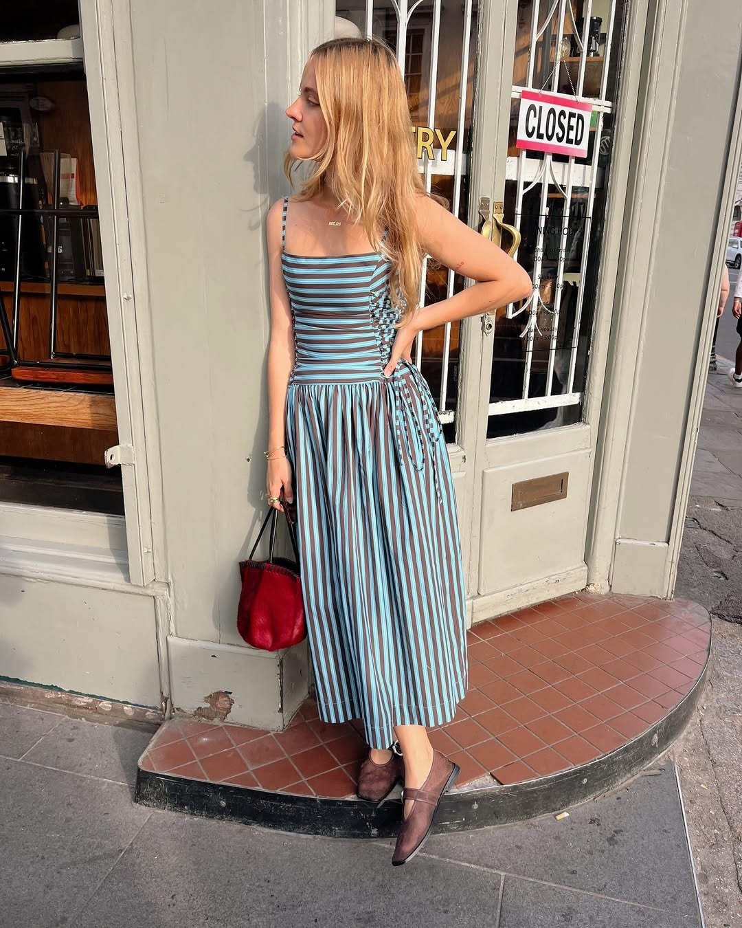 Mulher com vestido listrado azul, cor de Sagitário para 2026, posa na calçada em frente a loja fechada, segurando bolsa.