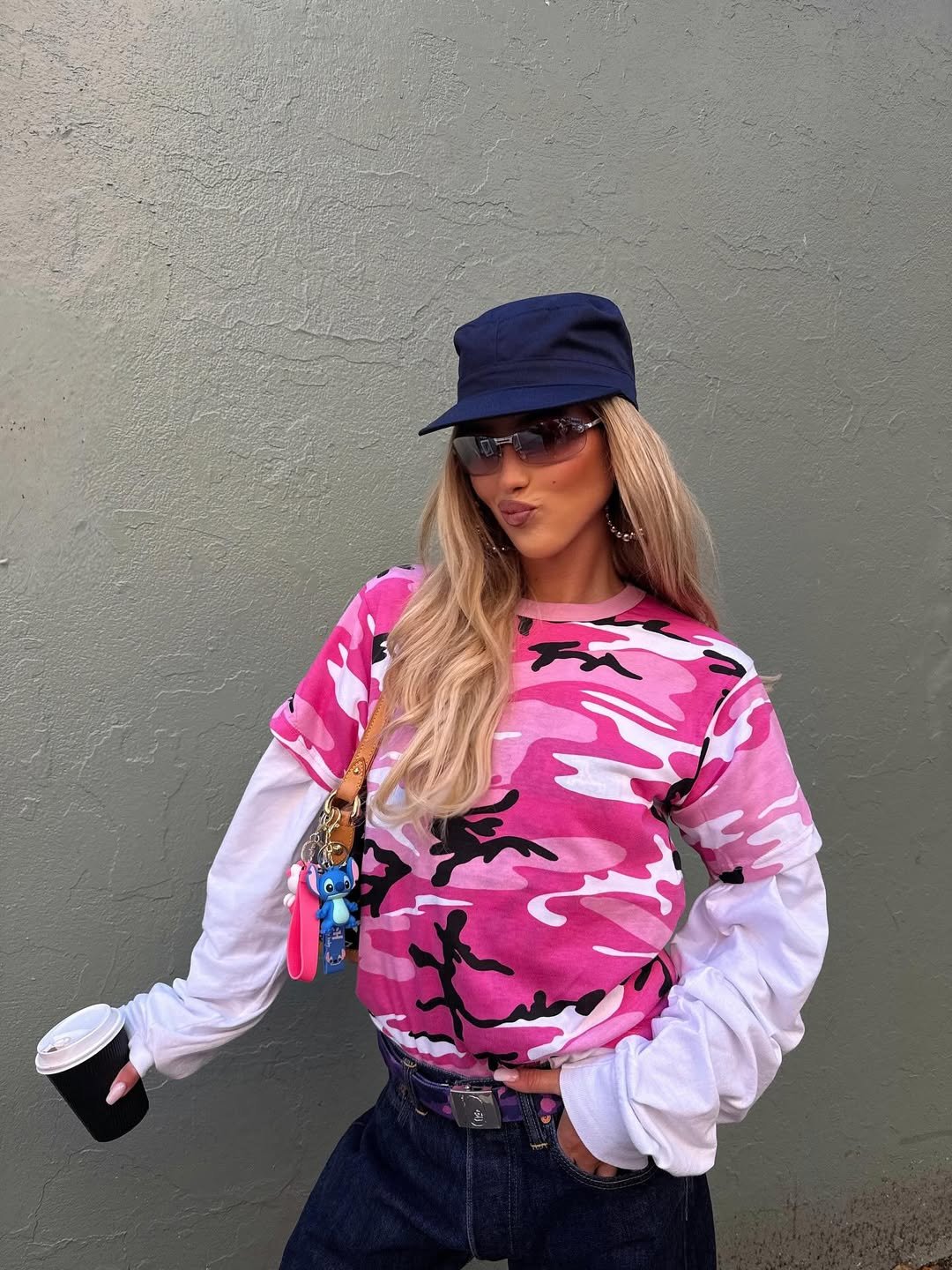 Mulher com blusa camuflada rosa e acessórios azuis, cores de Touro para 2026, posa com expressão confiante diante de parede