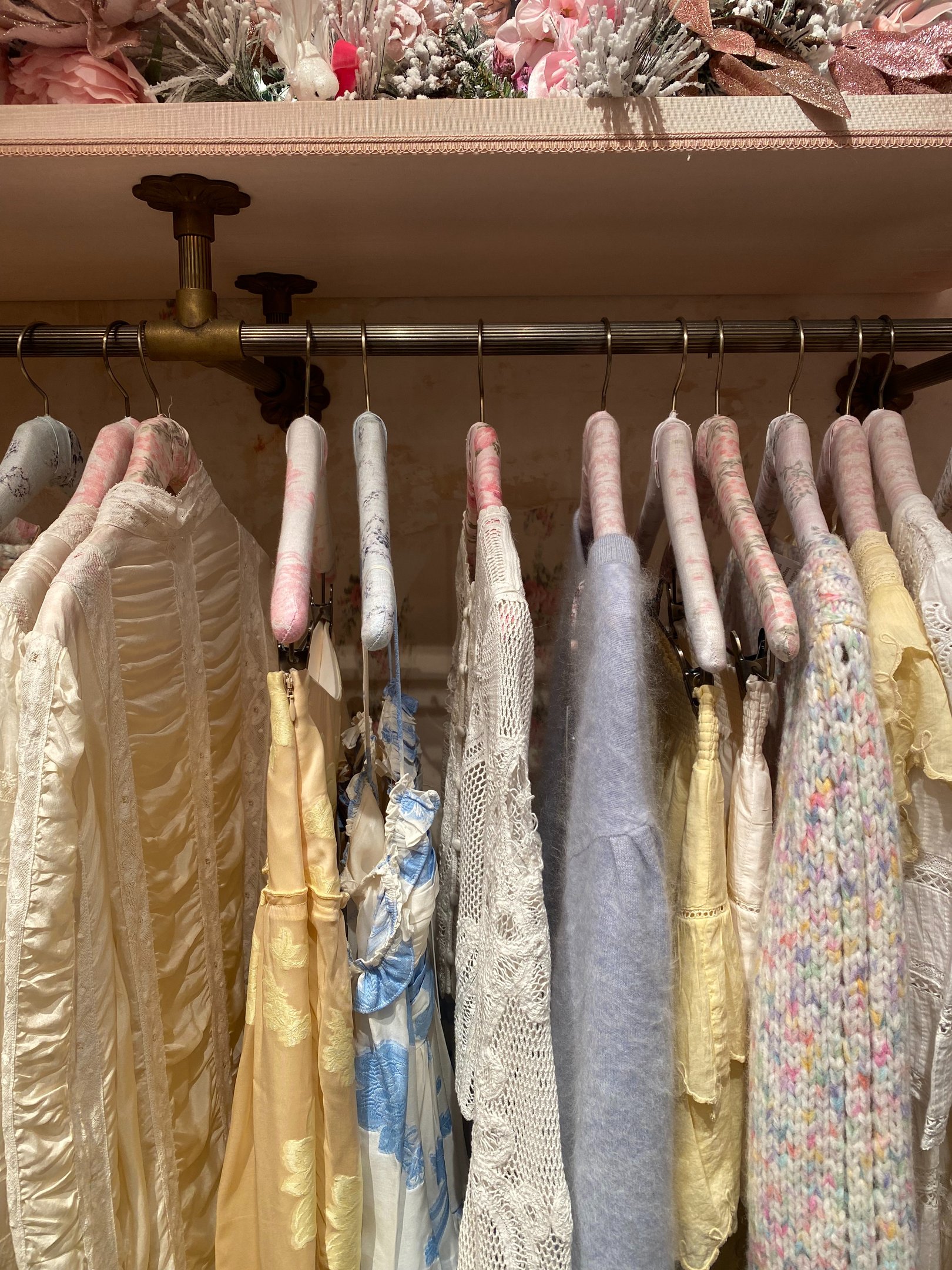 Closet pequeno com roupas delicadas em tons pastel, tecidos fluidos, crochê e tricô, criando atmosfera suave e feminina.