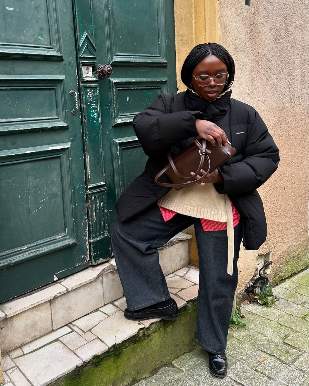 Modelo ajusta bolsa marrom enquanto veste jaqueta preta com gola funil acolchoada, suéter bege e calça jeans ampla, em clima
