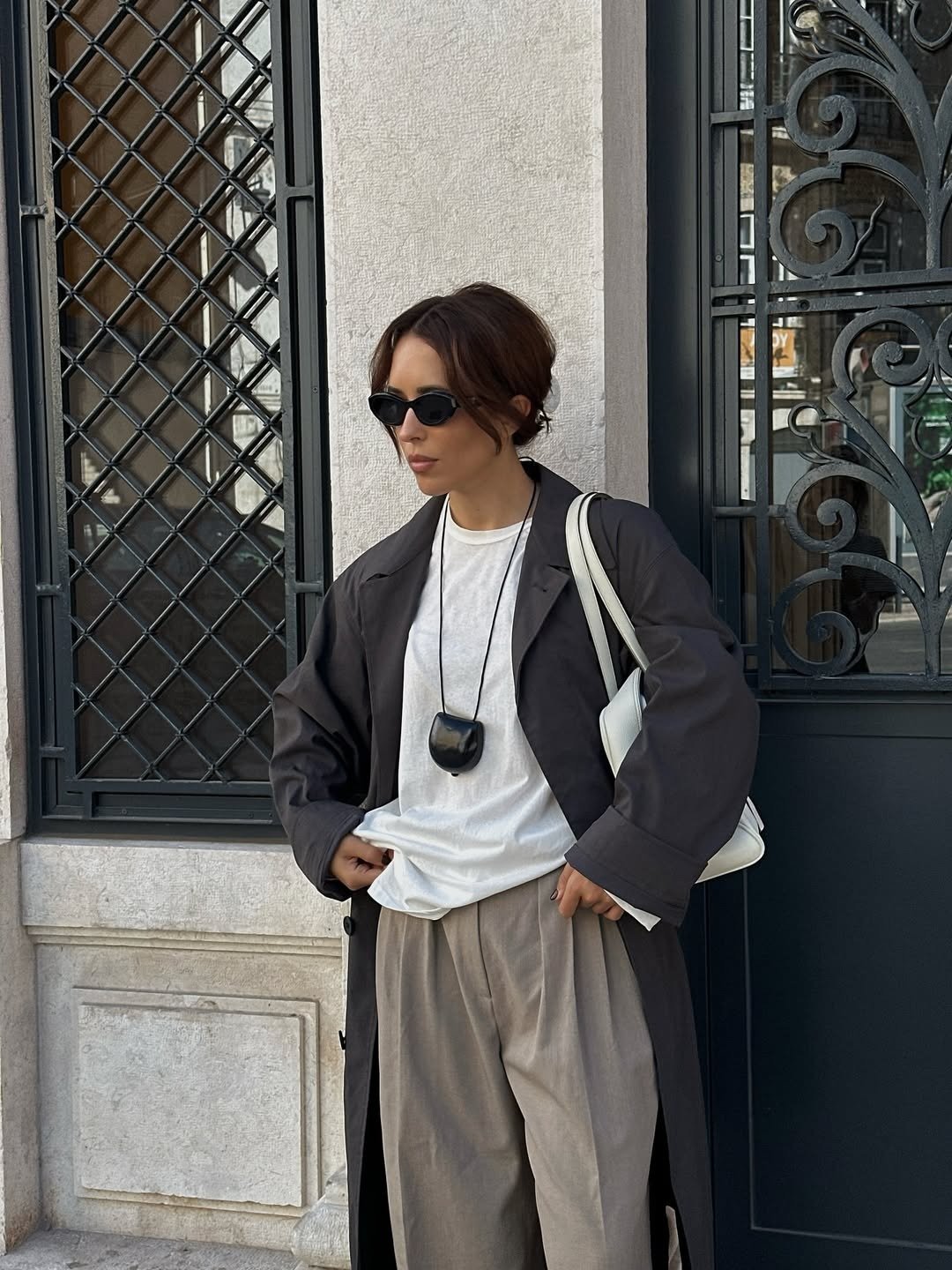 Modelo posa com colar funcional preto em formato arredondado, combinando com casaco longo cinza escuro, camiseta branca e