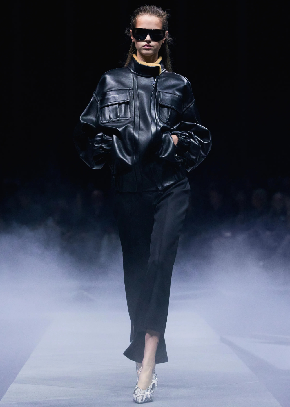 Modelo caminha com postura confiante usando jaqueta preta de couro com gola funil estruturada, calça reta preta e scarpin