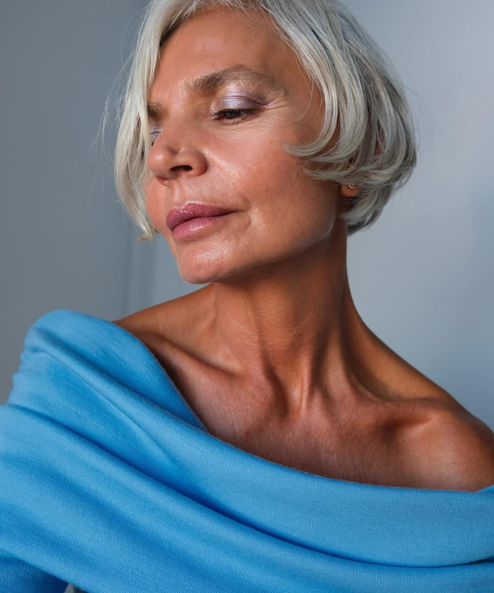 Mulher madura com pele radiante e maquiagem suave, veste blusa azul de tecido macio, expressando serenidade e cuidado;