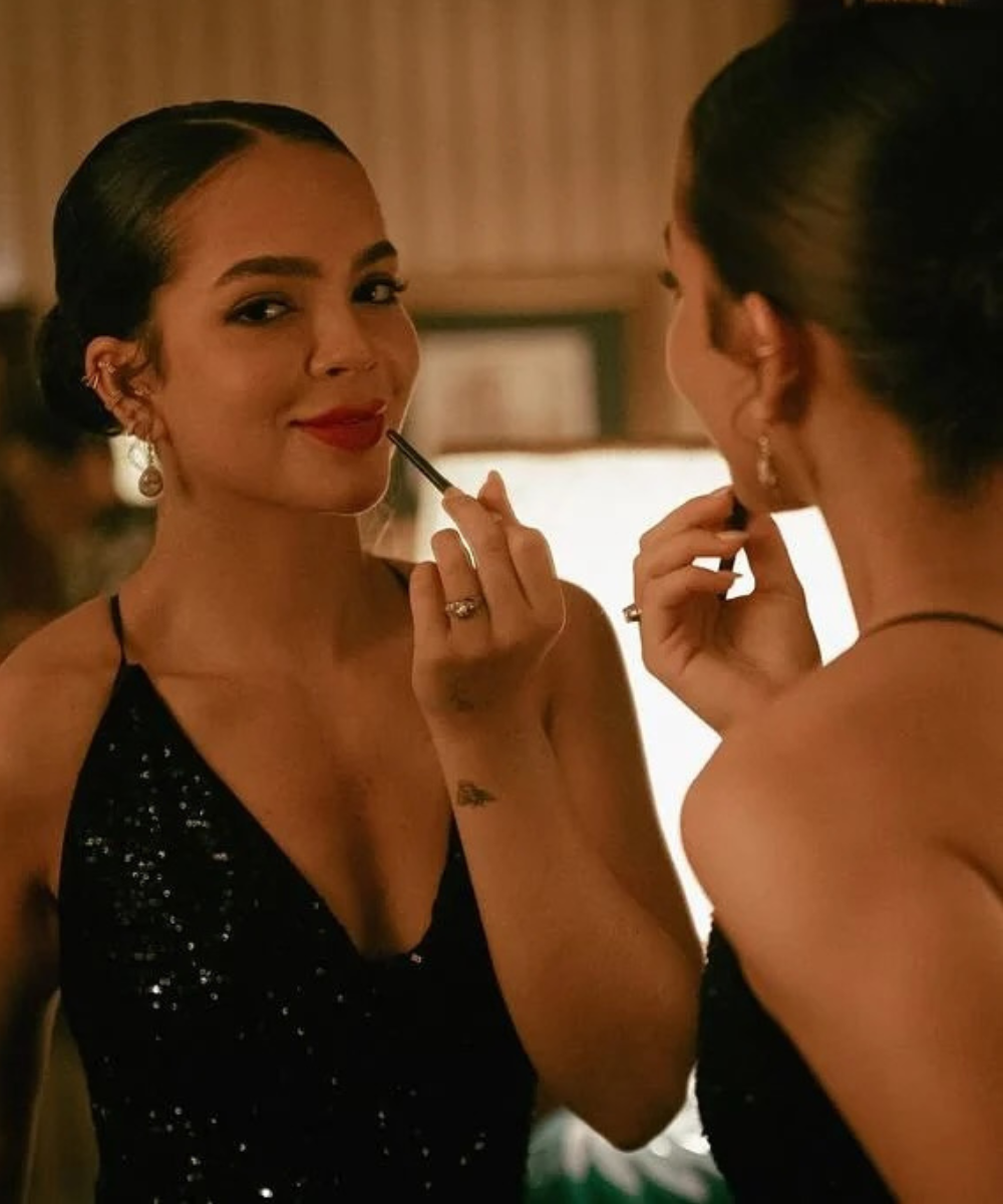 Mel Maia em Donos do Jogo aplica gloss vermelho vibrante, veste vestido preto de paetês com alças finas, em clima elegante e