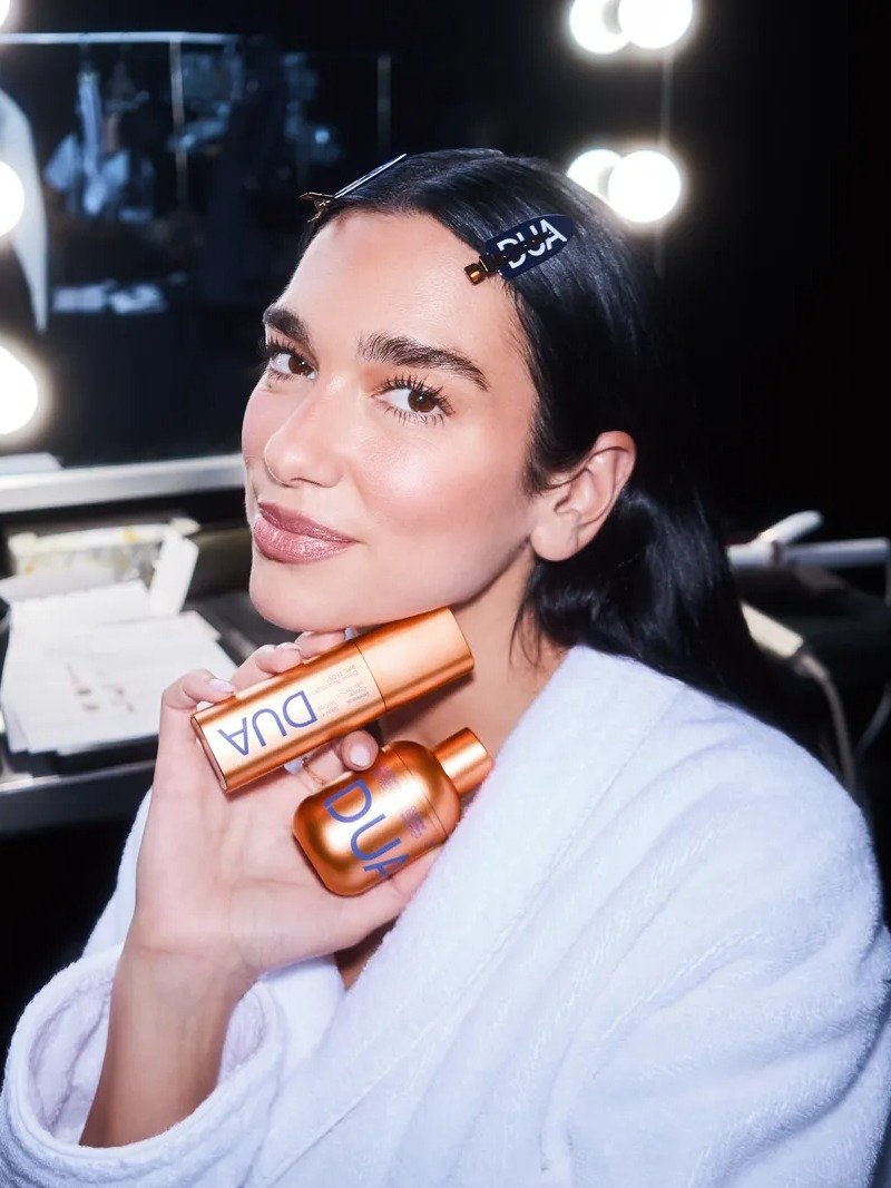 Dua Lipa exibe um look sofisticado e natural, com pele iluminada e lábios levemente glossados. Ela segura produtos de beleza em embalagens metálicas laranjas, destacando seu nome em azul.