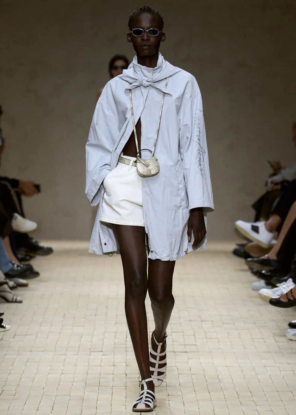 Mulher desfilando na passarela com short branco, camisa azul e sandália prata.