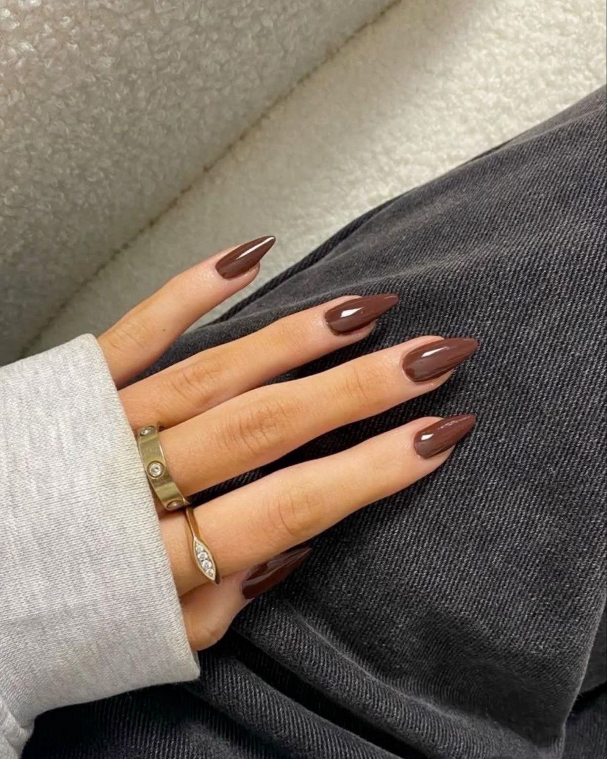 Unhas longas em formato stiletto com cores de esmalte 2026 em marrom brilhante, combinadas a anéis dourados e look casual