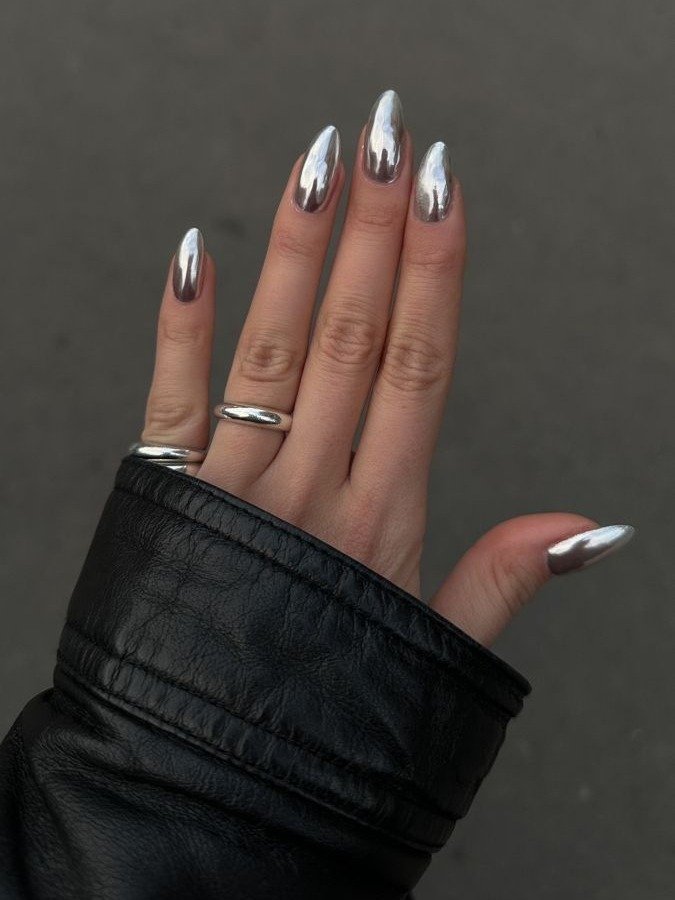 Unhas longas com esmalte cromado prata refletem as cores de esmalte 2026 em contraste com jaqueta de couro preta e anéis.