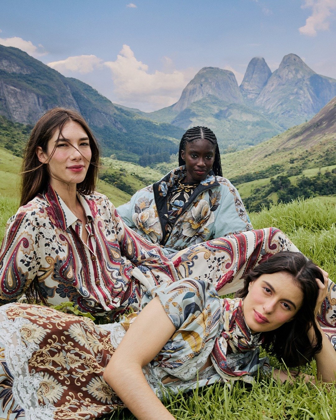 Três modelos posam relaxadas em paisagem montanhosa usando peças da Farm Global em tons terrosos e azuis da coleção