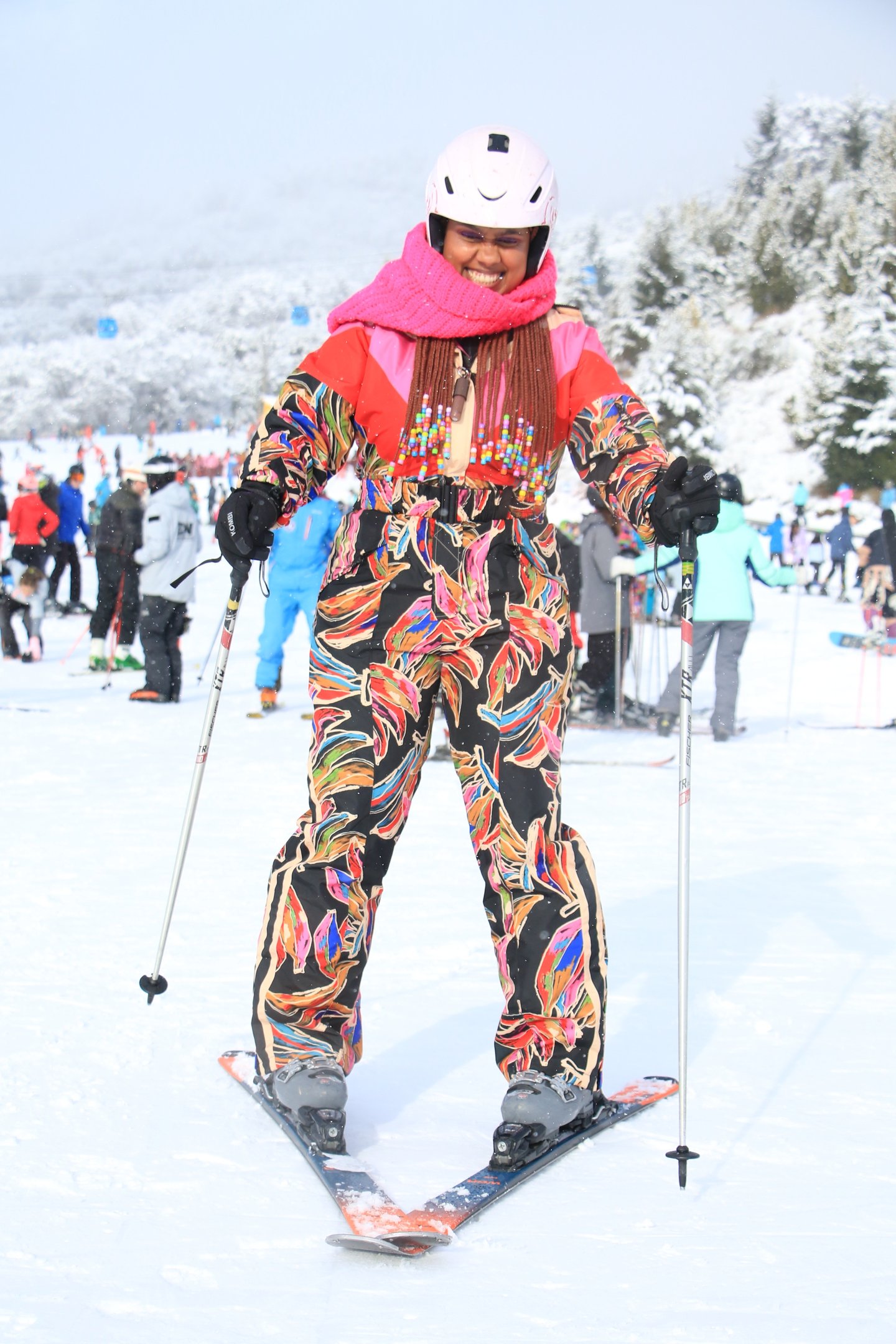 Modelo sorri e esquia vestindo macacão estampado colorido Farm Global, cachecol rosa vibrante e capacete branco, em cenário