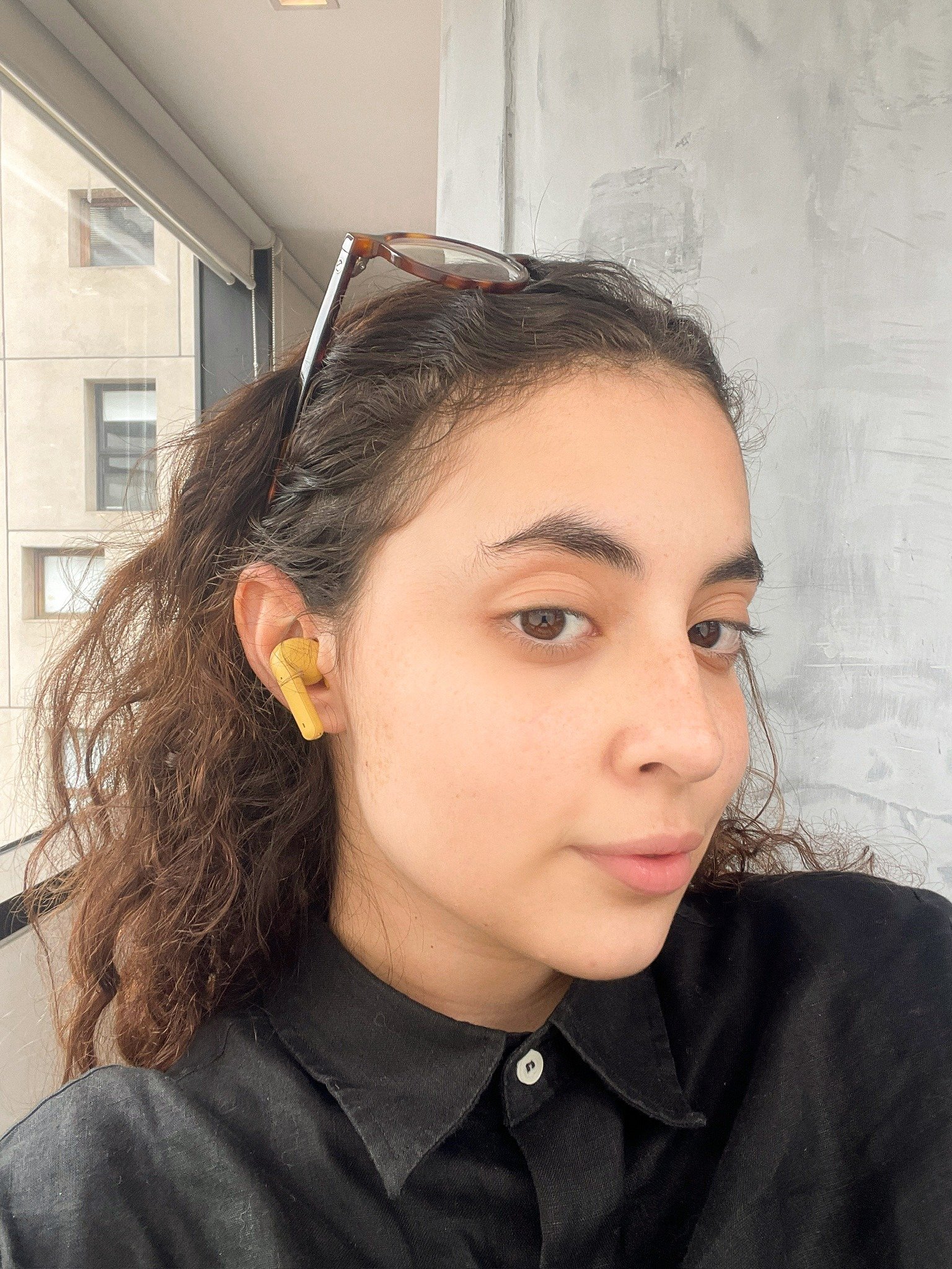 A imagem apresenta uma jovem com cabelos cacheados, usando um fone de ouvido da Farm na cor amarela, que se destaca no contexto de moda e beleza. Ela veste uma camisa preta de gola, que contrasta com o tom vibrante do acessório.