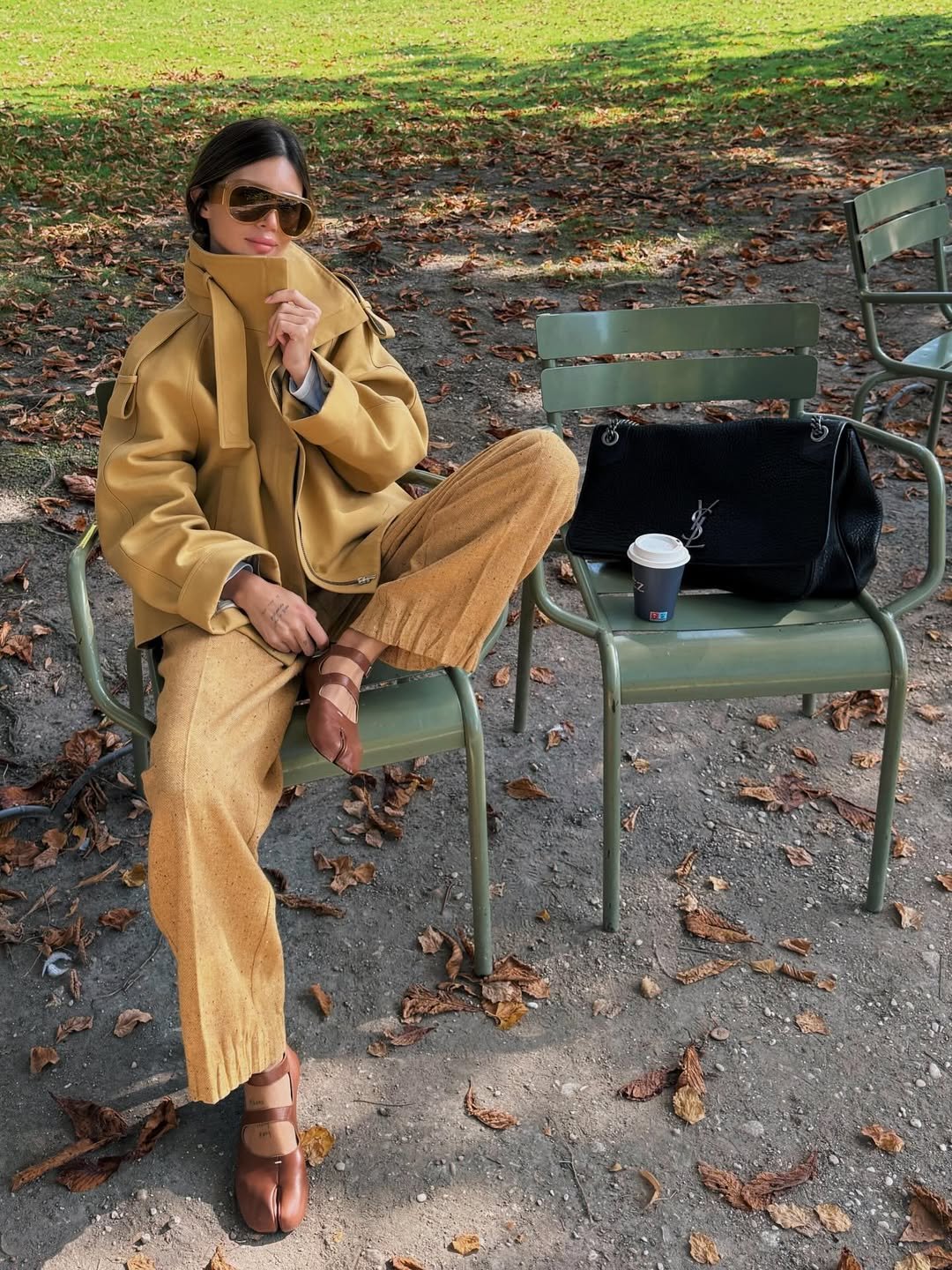 Modelo senta casualmente em cadeira verde ao ar livre, vestindo casaco mostarda com gola funil estruturada e calça ampla