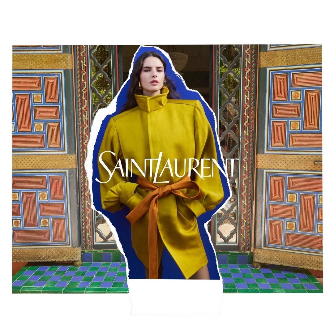 Modelo posa com casaco mostarda de tecido estruturado, gola funil alta e cinto marrom amarrado, em cenário com portas