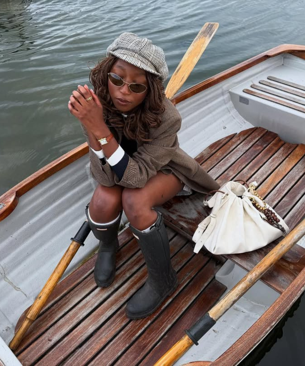 Mulher sentada em um barco no rio, usando boina e óculos de sol. Usa galochas bonitas pretas.