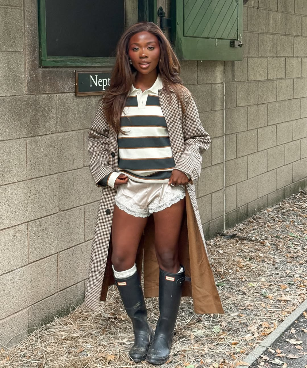 Mulher em pé usando blusa polo listrada verde, casaco longo, shorts de cetim e galochas bonitas.