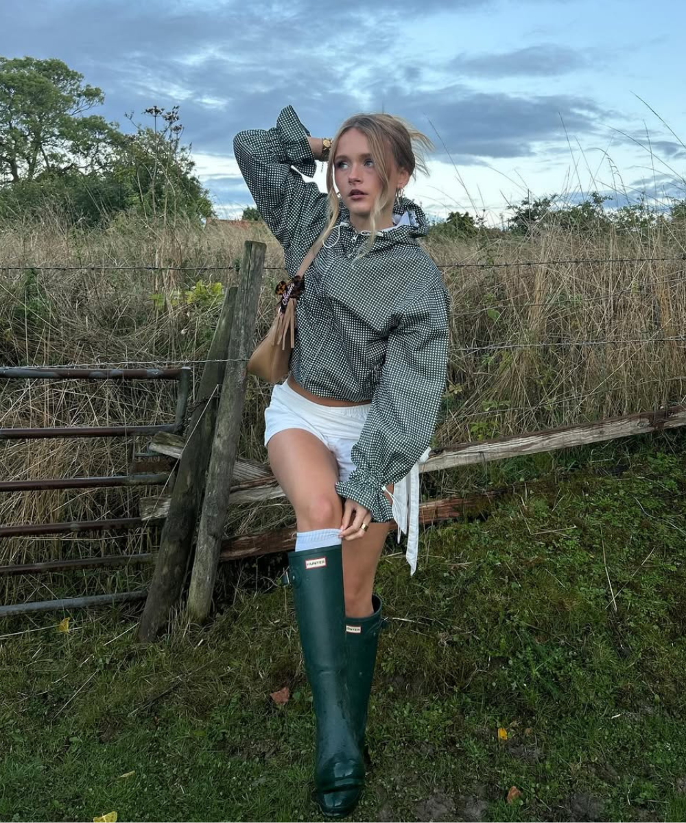 Mulher em pé no meio do campo usando saia de renda, jaqueta tipo funnel verde e galochas verdes.