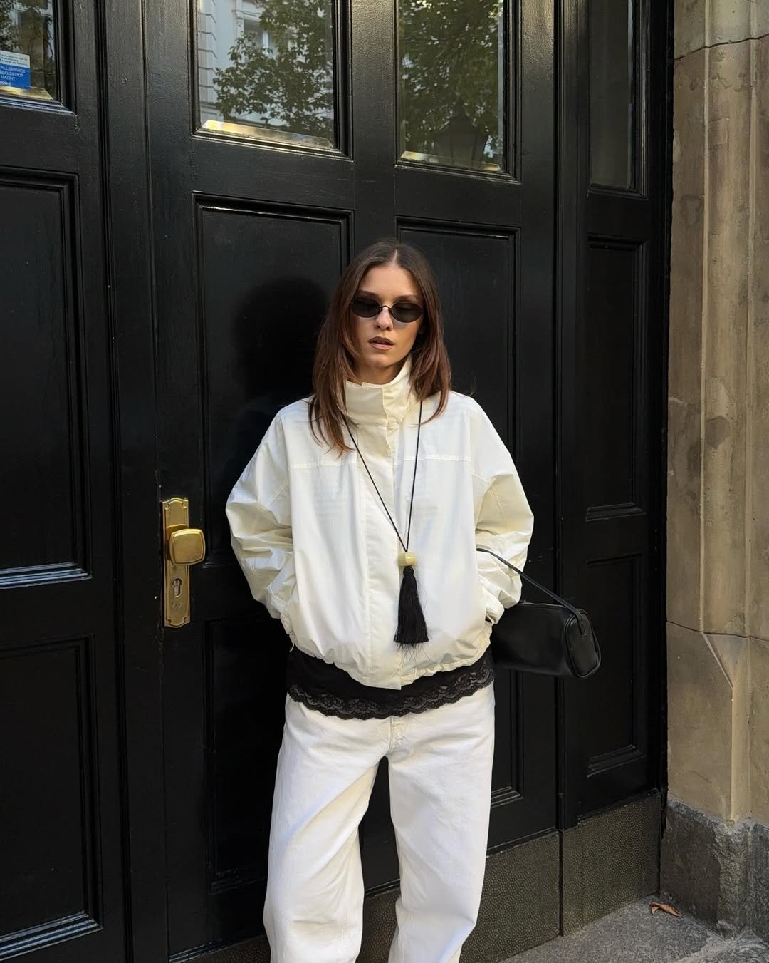Modelo posa com jaqueta branca de tecido leve e gola funil alta, combinada a calça branca e acessórios pretos, em clima