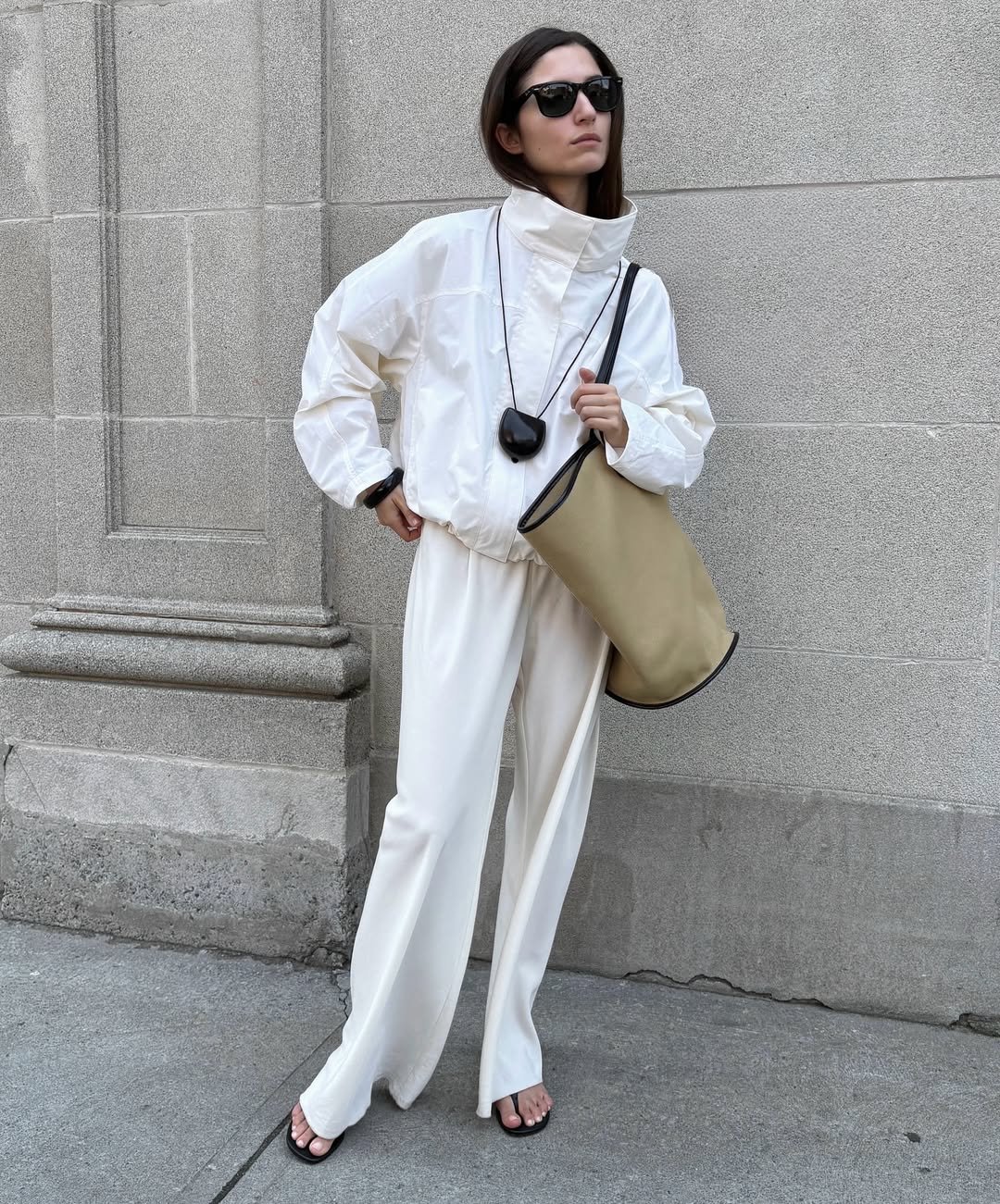 Modelo posa com jaqueta branca de gola funil estruturada, calça ampla e bolsa bege, em look minimalista e sofisticado.