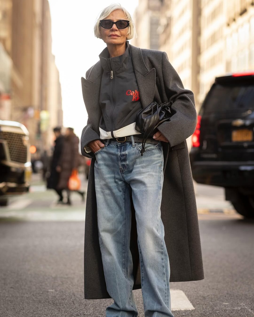 Modelo posa com jaqueta cinza de gola funil em tecido técnico, casaco longo cinza e jeans claro, criando look urbano