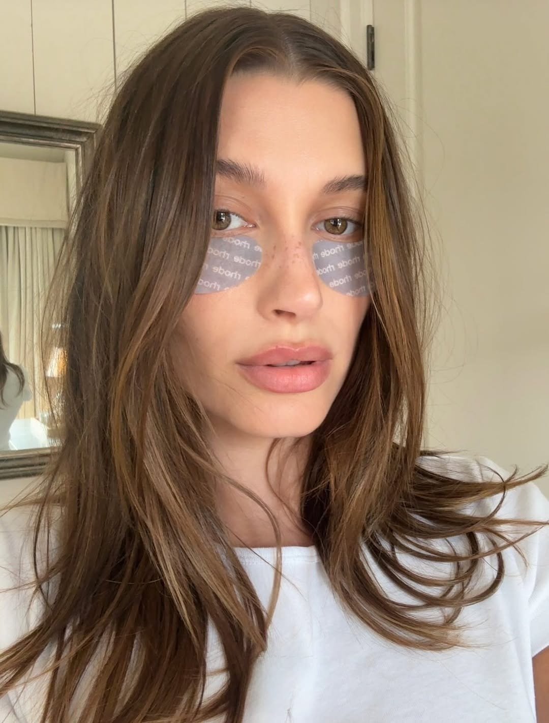 Modelo posa com olhar sereno e natural, usando patches PRP sob os olhos, cabelo solto e camiseta branca em ambiente claro.