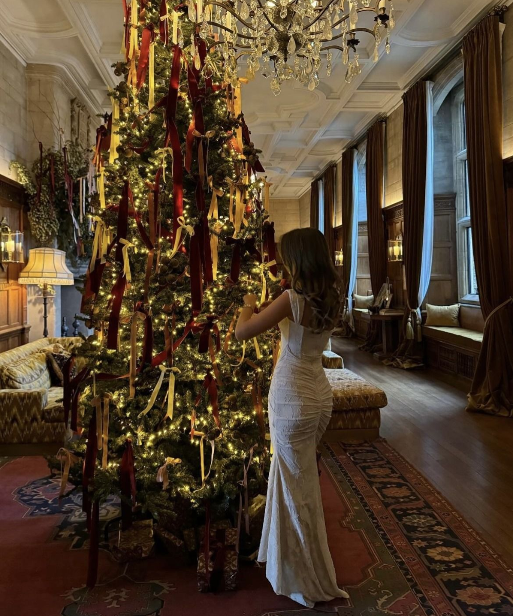 Modelo veste vestido longo branco justo de tecido texturizado e decote quadrado, decorando árvore de Natal com fitas