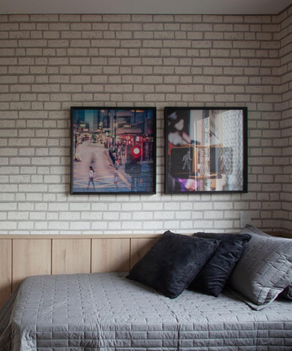 Quarto com papéis de parede que imitam tijolos claros, cama com colcha cinza texturizada e almofadas pretas, ambiente