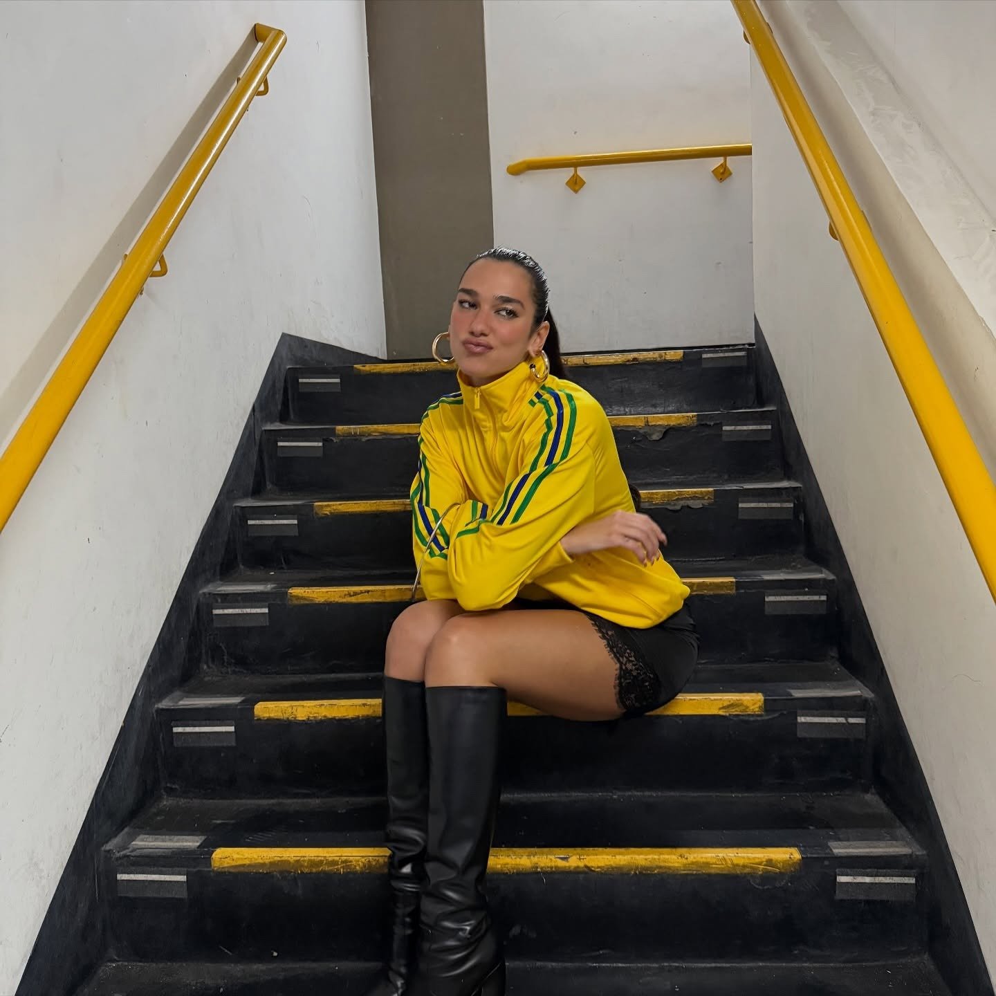 Modelo senta na escada com jaqueta da Dua Lipa amarela vibrante, listras verdes, combinada a shorts pretos e botas longas,