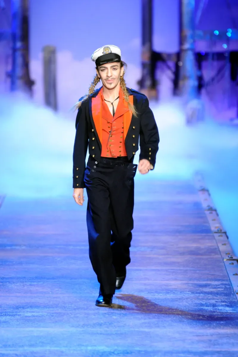 Modelo caminha confiante com look inspirado em uniforme naval, casaco preto com botões dourados e colete laranja vibrante,