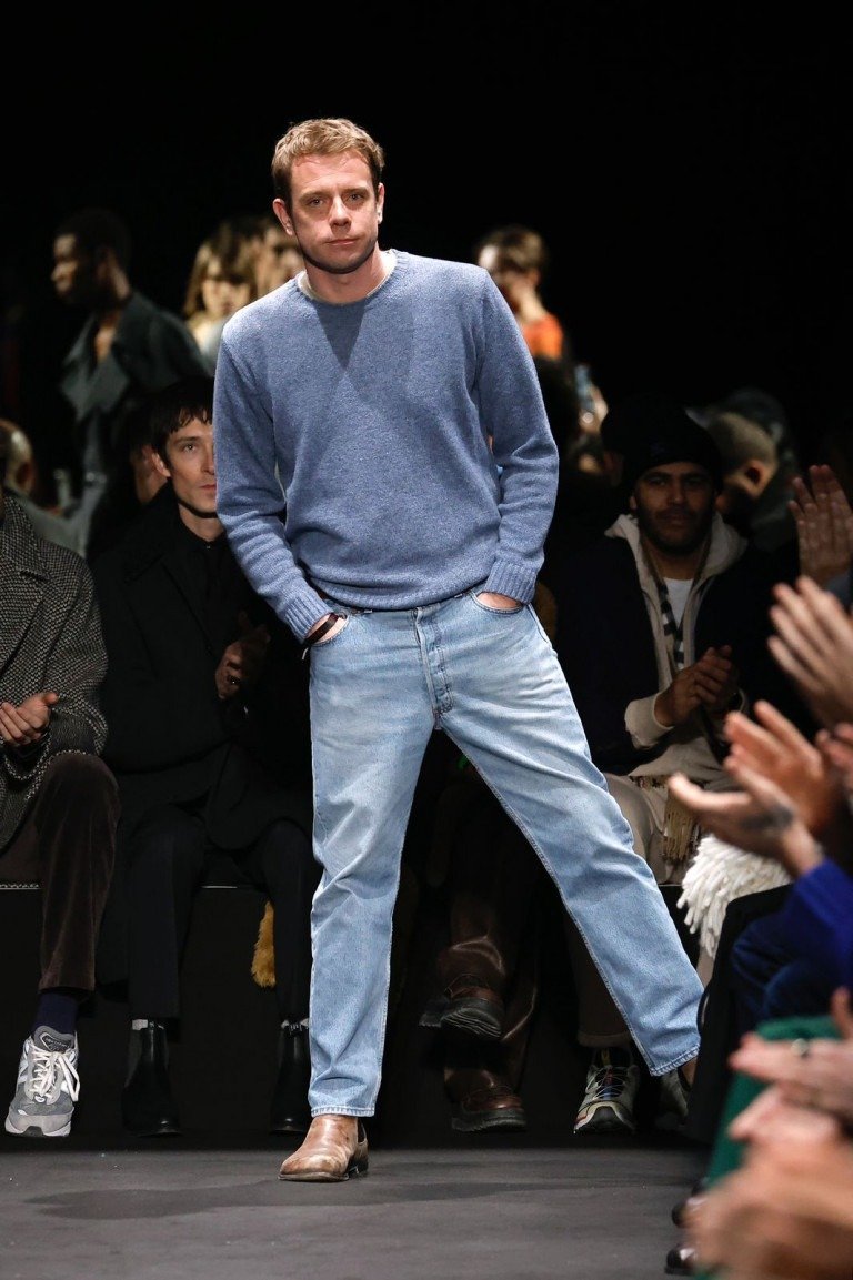 Diretor criativo posa confiante com suéter azul de malha leve, jeans claro de corte reto e botas de couro marrom, em clima