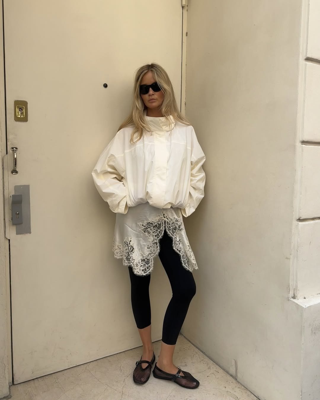 Modelo posa com blusa off-white de gola funil ampla em tecido estruturado, saia com renda delicada e legging preta, em