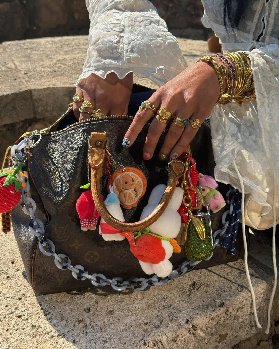 Mãos adornadas com anéis dourados e pulseiras combinam com bolsa cravejada de chaveiros coloridos de kidults.