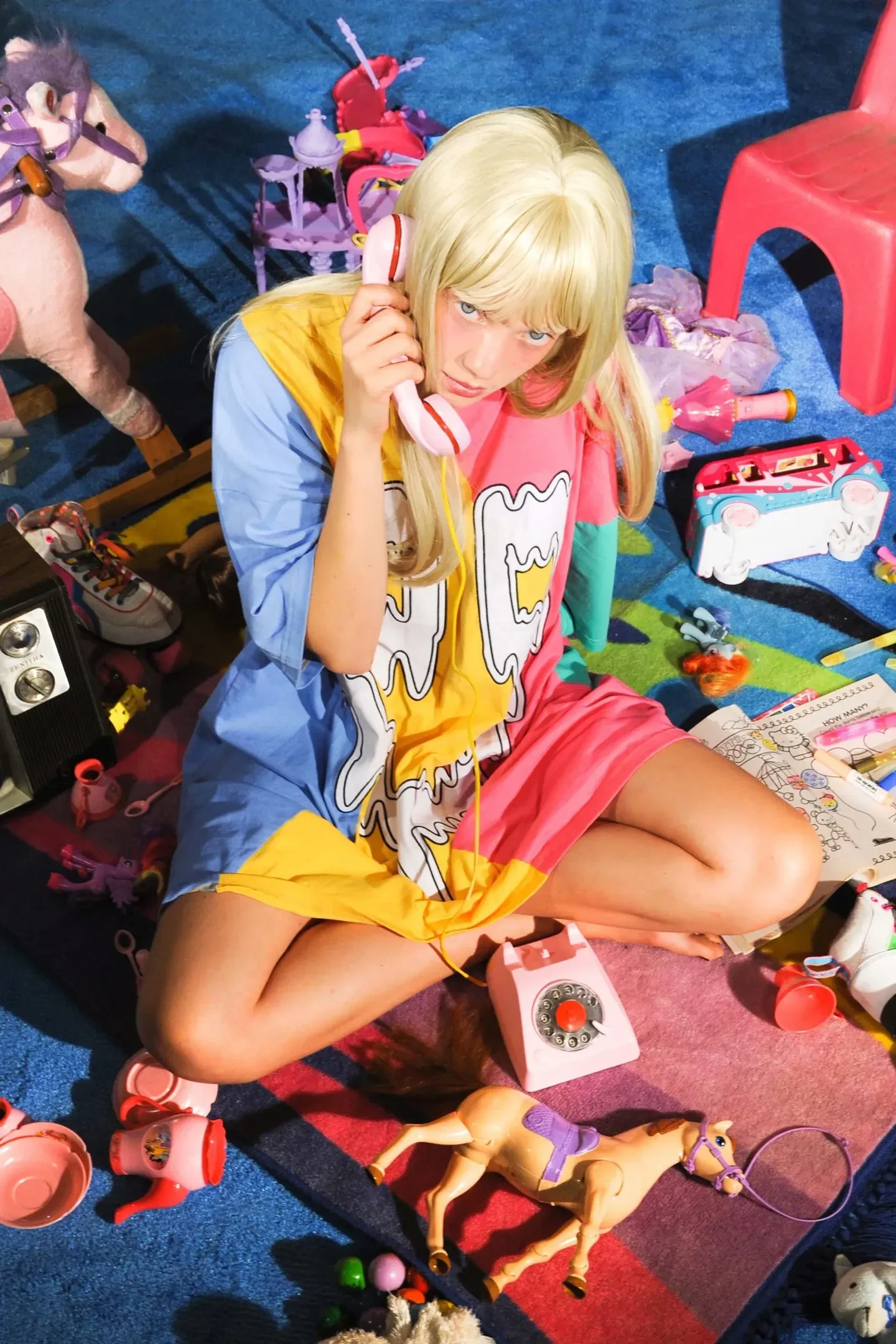Modelo senta no chão colorido cercada por brinquedos, vestindo vestido oversized, representando kidults.