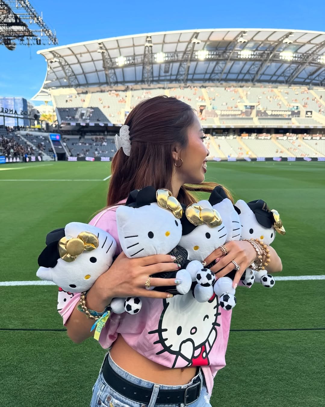 Jovem kidult posa sorrindo no estádio com camiseta rosa de Hello Kitty e abraça quatro pelúcias da personagem com laços.