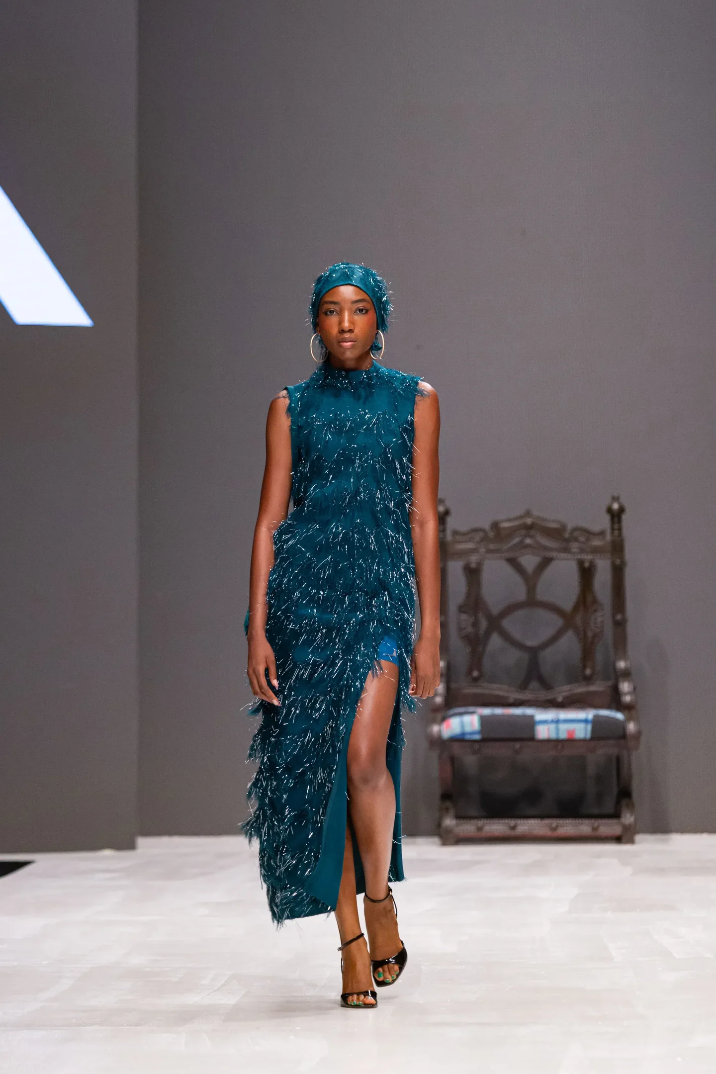 Durante a Lagos Fashion Week, uma modelo desfila com um vestido longo na cor verde-água. A peça, com detalhes de franjas, apresenta uma fenda lateral que destaca a silhueta. A modelo usa um lenço combinando e brincos grandes, complementando o visual.