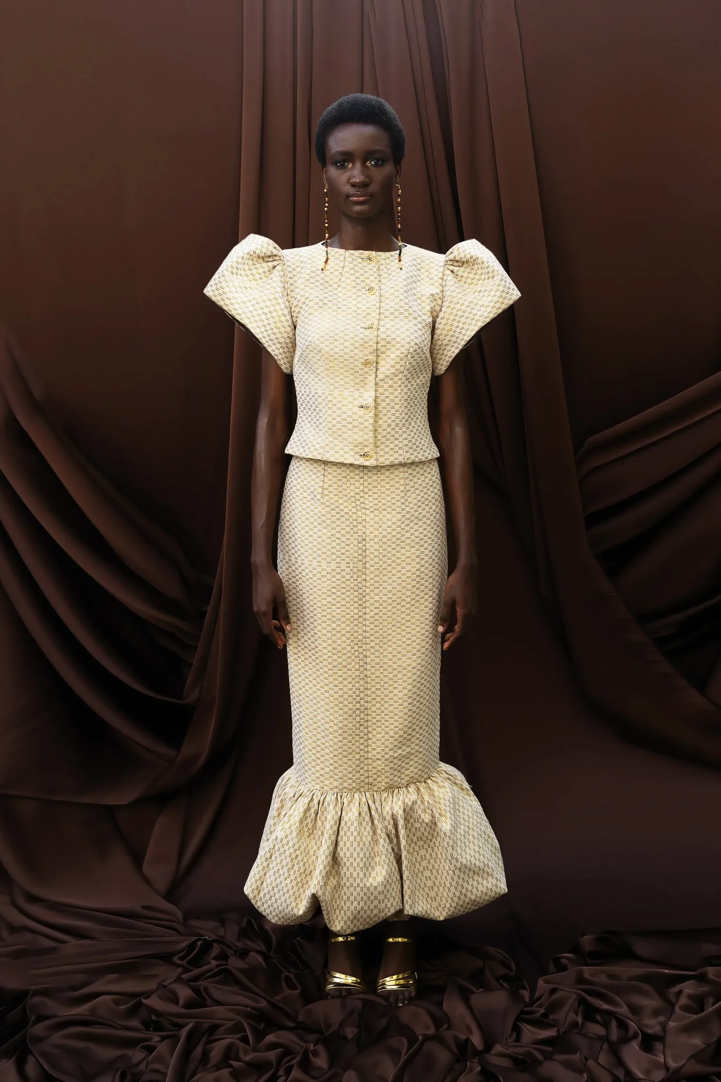 Durante a Lagos Fashion Week, destaca-se um vestido elegante em tom claro com mangas volumosas e detalhes em textura. A peça, que termina em babados, é complementada por acessórios dourados.