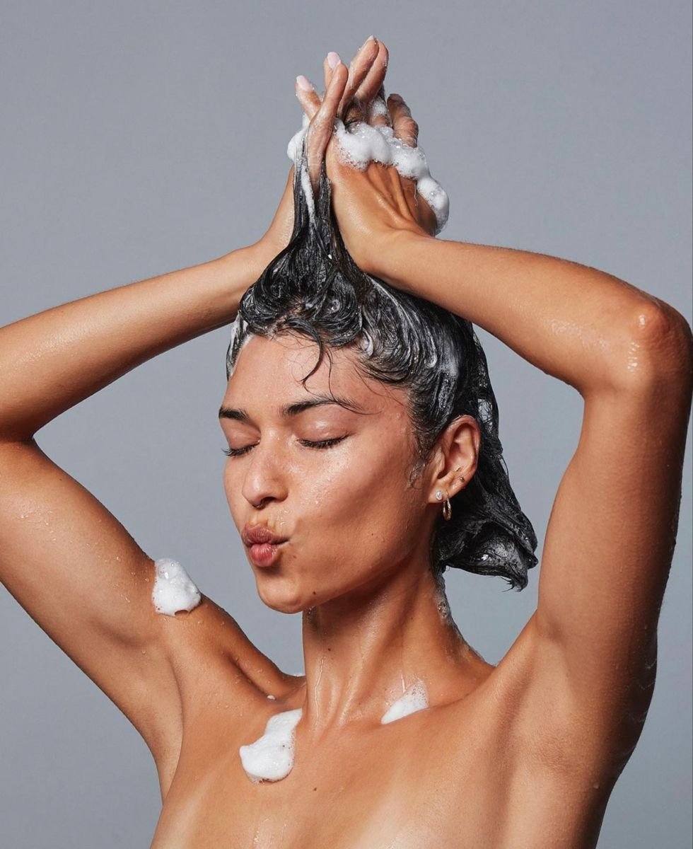 Mulher de pele bronzeada lava o cabelo com espuma branca, mostrando como lavar o cabelo corretamente.