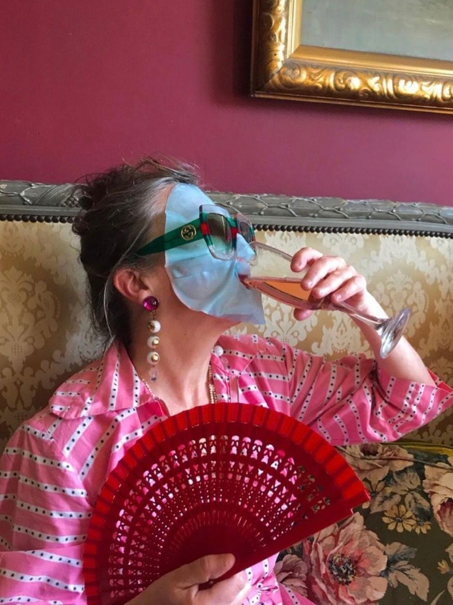 A imagem apresenta uma mulher relaxando em um ambiente elegante, segurando um leque vermelho vibrante. Ela usa um creme facial azul e óculos de sol estilizados, complementados por brincos de pérola.