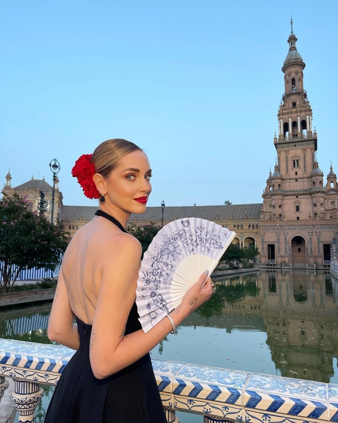 A imagem apresenta uma mulher elegante, usando um vestido preto com costas nuas, que destaca sua silhueta. Ela segura um leque branco decorado com desenhos florais, complementando seu visual.