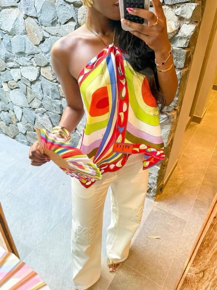 Uma mulher segura um leque colorido enquanto faz uma selfie. Ela veste uma blusa vibrante com estampas geométricas em tons de verde, vermelho e amarelo, combinada com calças brancas. O ambiente tem paredes de pedra, criando um contraste elegante.