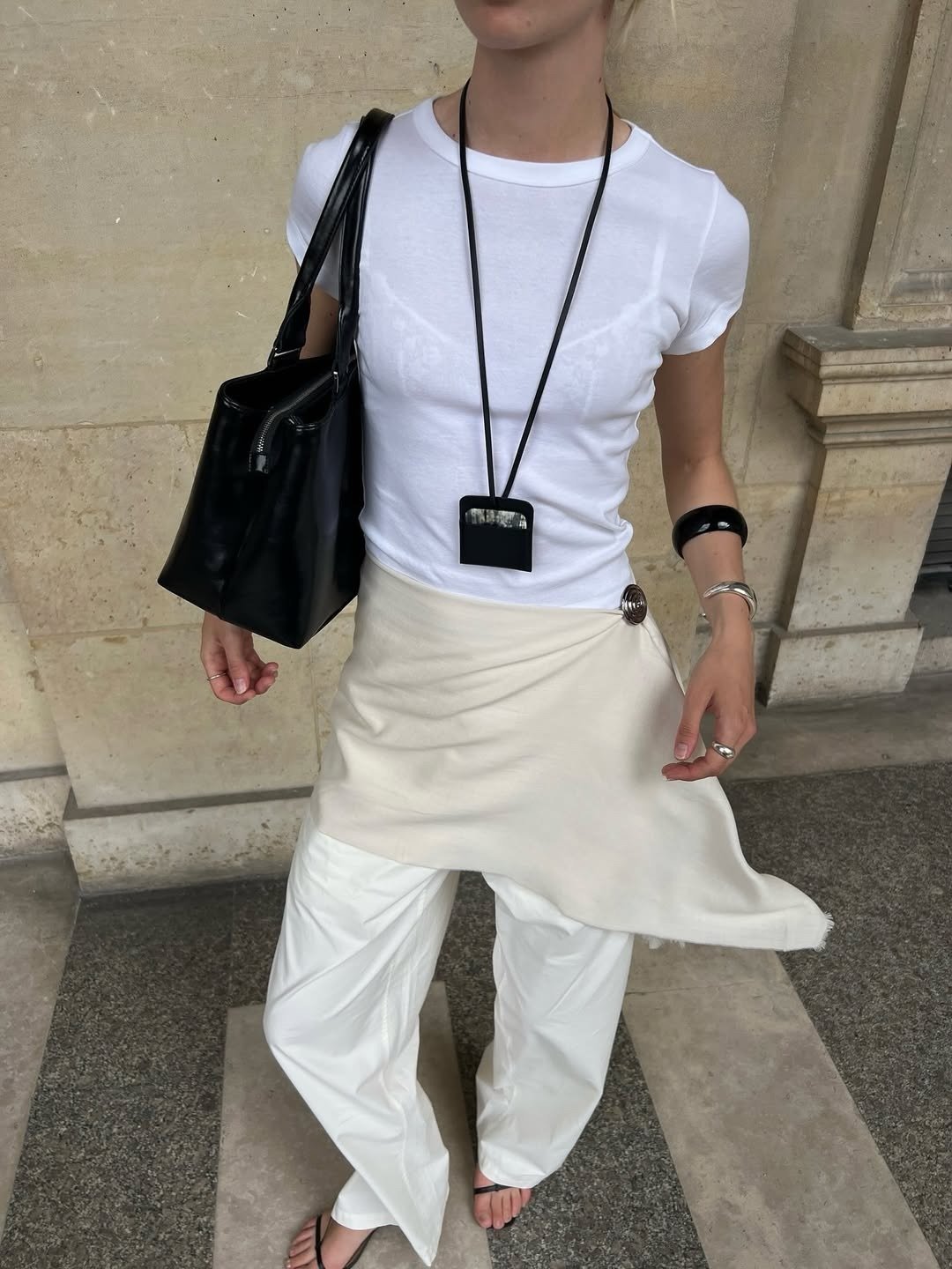 Modelo posa com colar funcional preto de design minimalista sobre camiseta branca justa, saia bege assimétrica e calça ampla