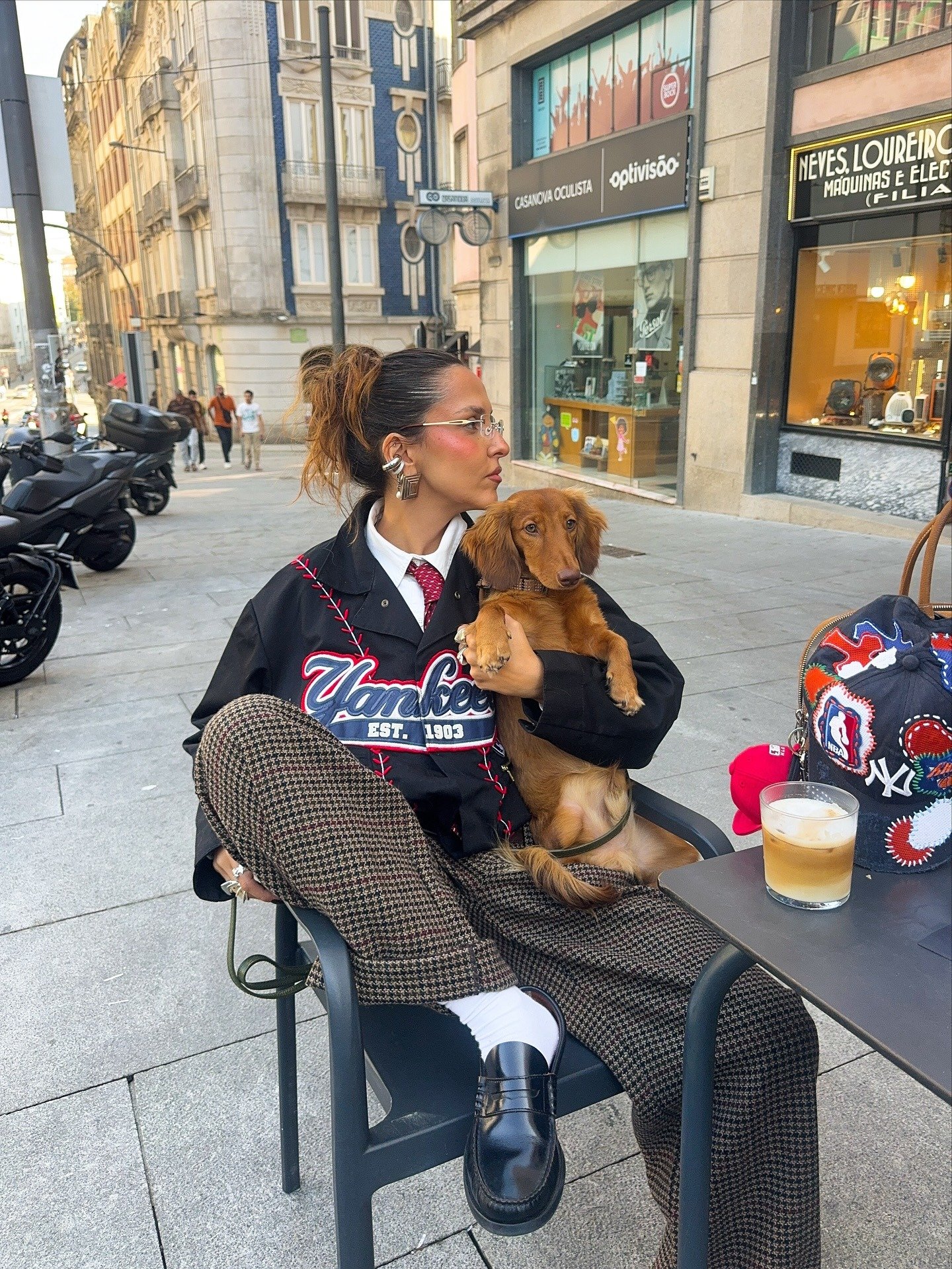 O look apresenta uma combinação estilosa com loafer de couro preto, um clássico que adiciona sofisticação ao visual. A pessoa usa uma blusa oversized com estampa, calças em padrão xadrez e acessórios modernos, como brincos grandes e óculos.