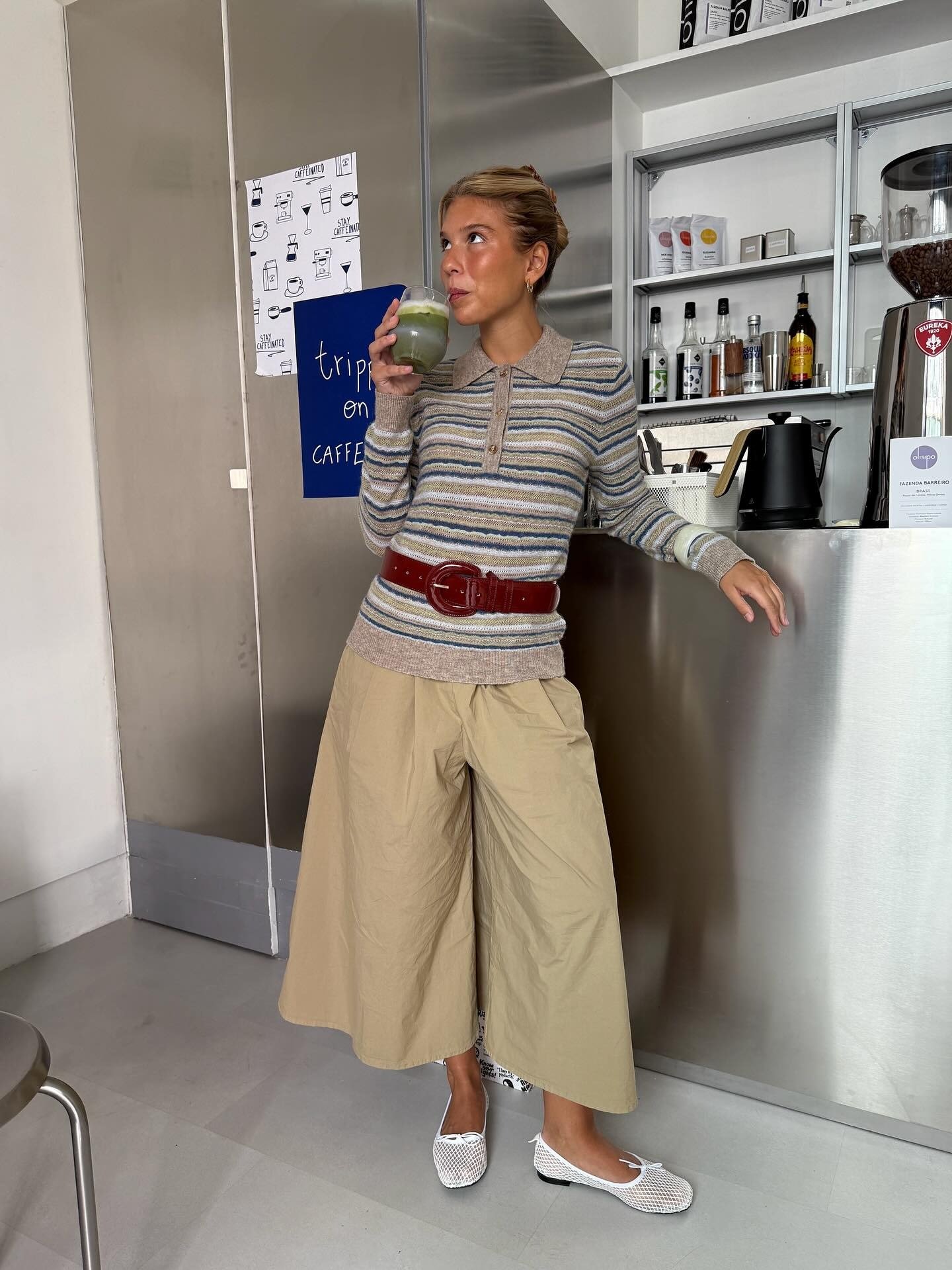 Look com pantacourt bege de tecido leve combinado a suéter listrado em tons neutros e cinto vinho, modelo posa bebendo suco.
